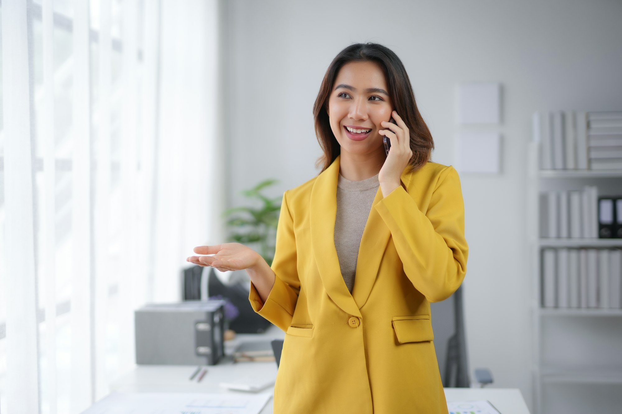 Businesswoman in yellow blazer talking on phone, standing in modern office